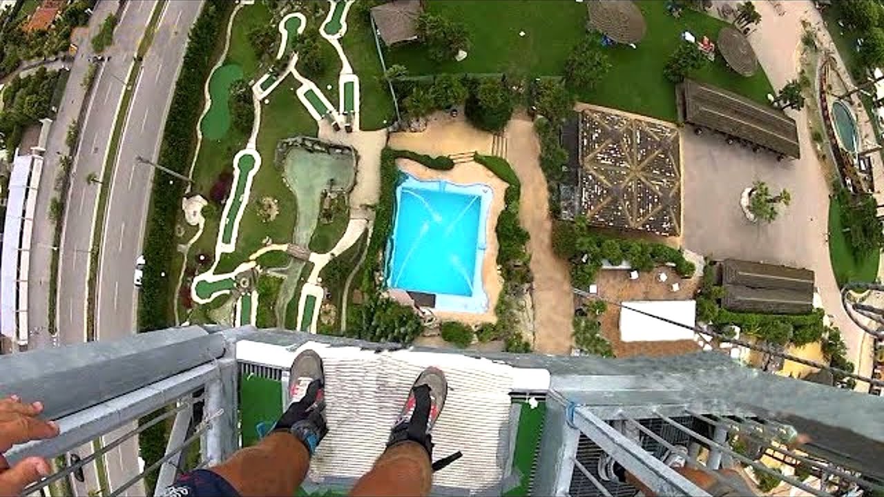 Waghalsiger Cliff-Diver beim Absprung von hoher Klippe Ein wagemutiger Cliff-Diver stürzt sich von einer hohen Felsklippe in kristallklares Wasser; Luftaufnahme zeigt die beeindruckende Höhe und den kraftvollen Aufprall.