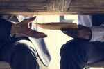 Zwei Männer im Anzug beim Austausch eines Umschlags unter dem Tisch – Symbol für intransparente politische Praktiken.